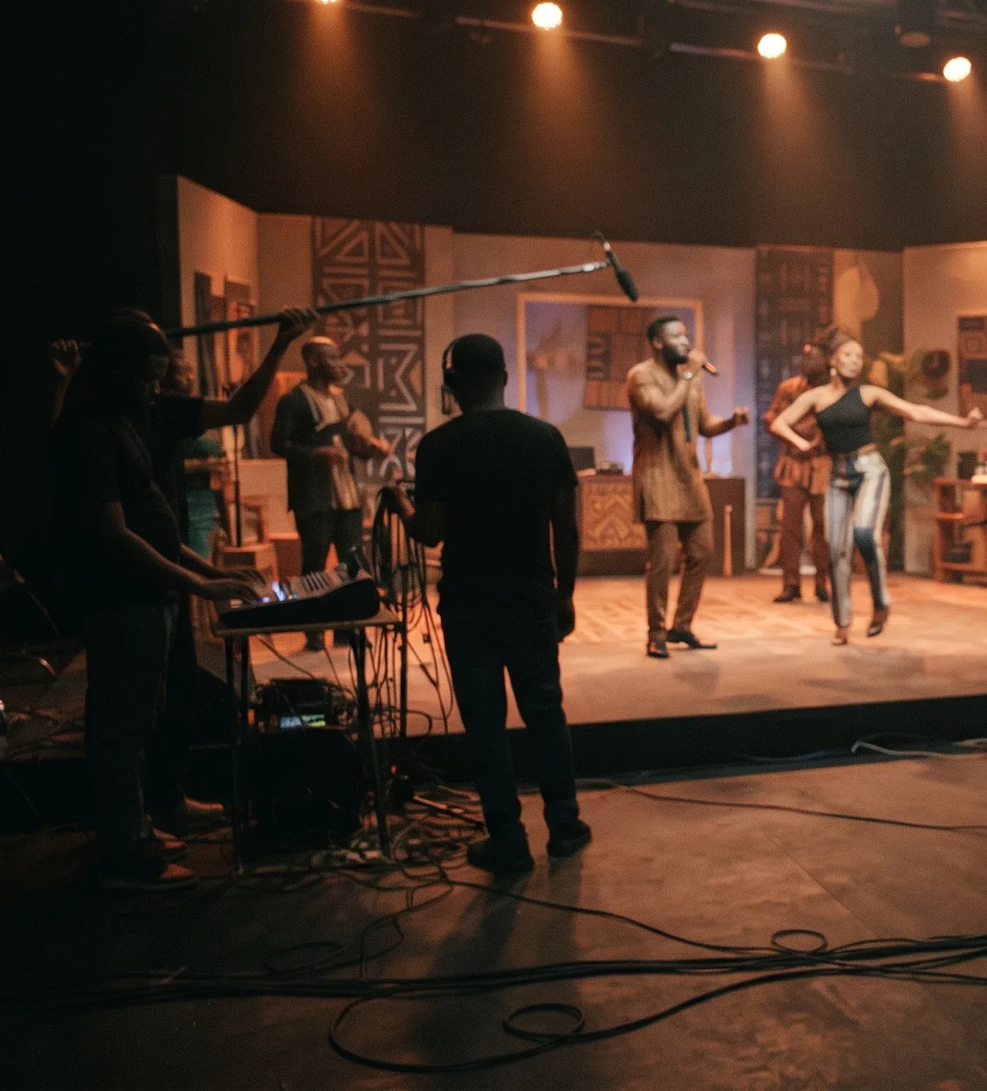 A dimly lit studio stage with two performers under warm spotlights, crew members with a boom microphone and a keyboard in the foreground.