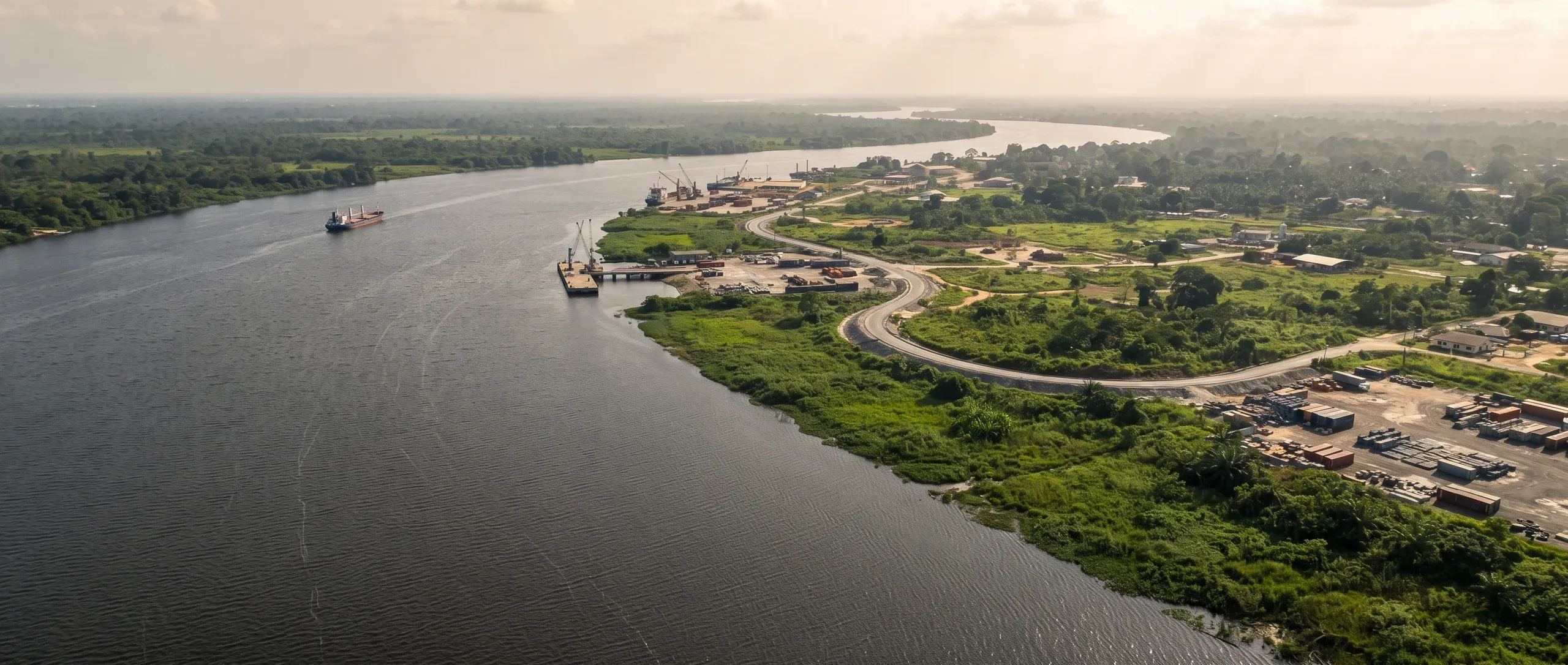 Aerial view of a broad river bending past a small riverside port, with a road winding inland through green land.